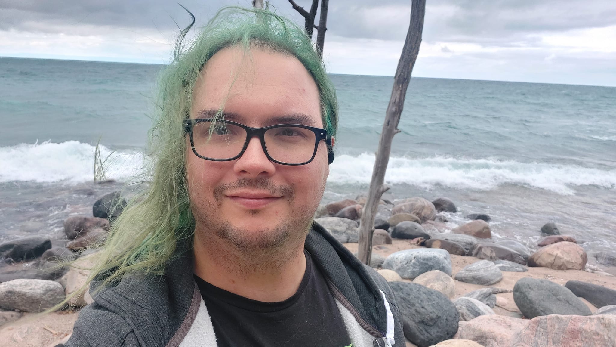 A man with long green hair and no beard stands in front of Lake Huron, waves crashing on the breakwater, Photo 1